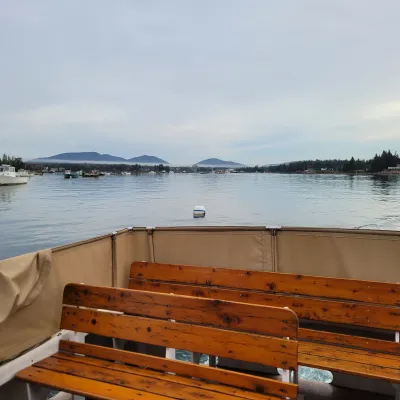 View of a calm lake from a boat deck with empty benches, boats, and mountains in the background.