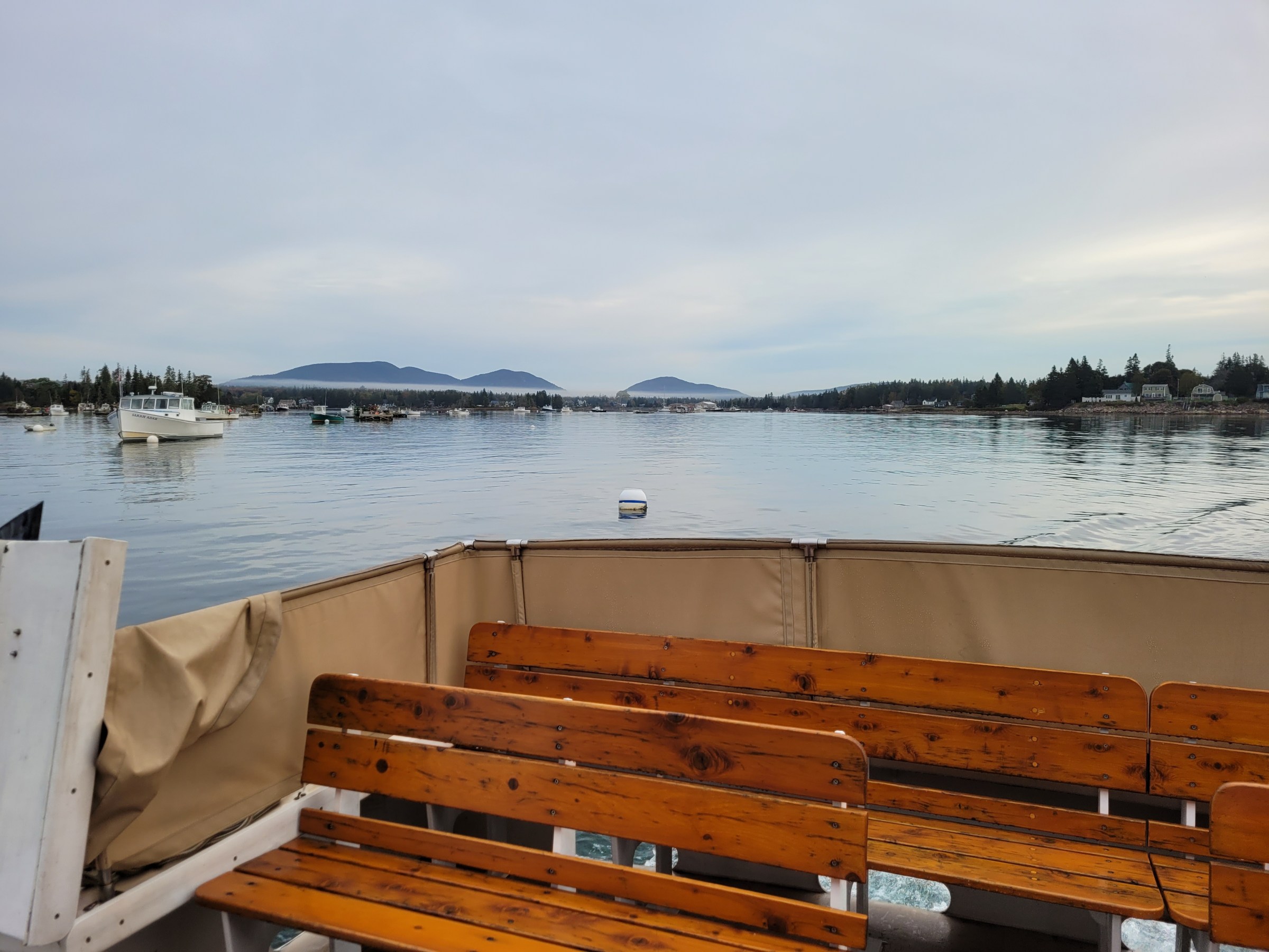View of a calm lake from a boat deck with empty benches, boats, and mountains in the background.