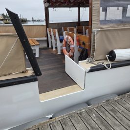 Boat with canvas cover, wooden deck, lifebuoy, docked beside wooden pier.