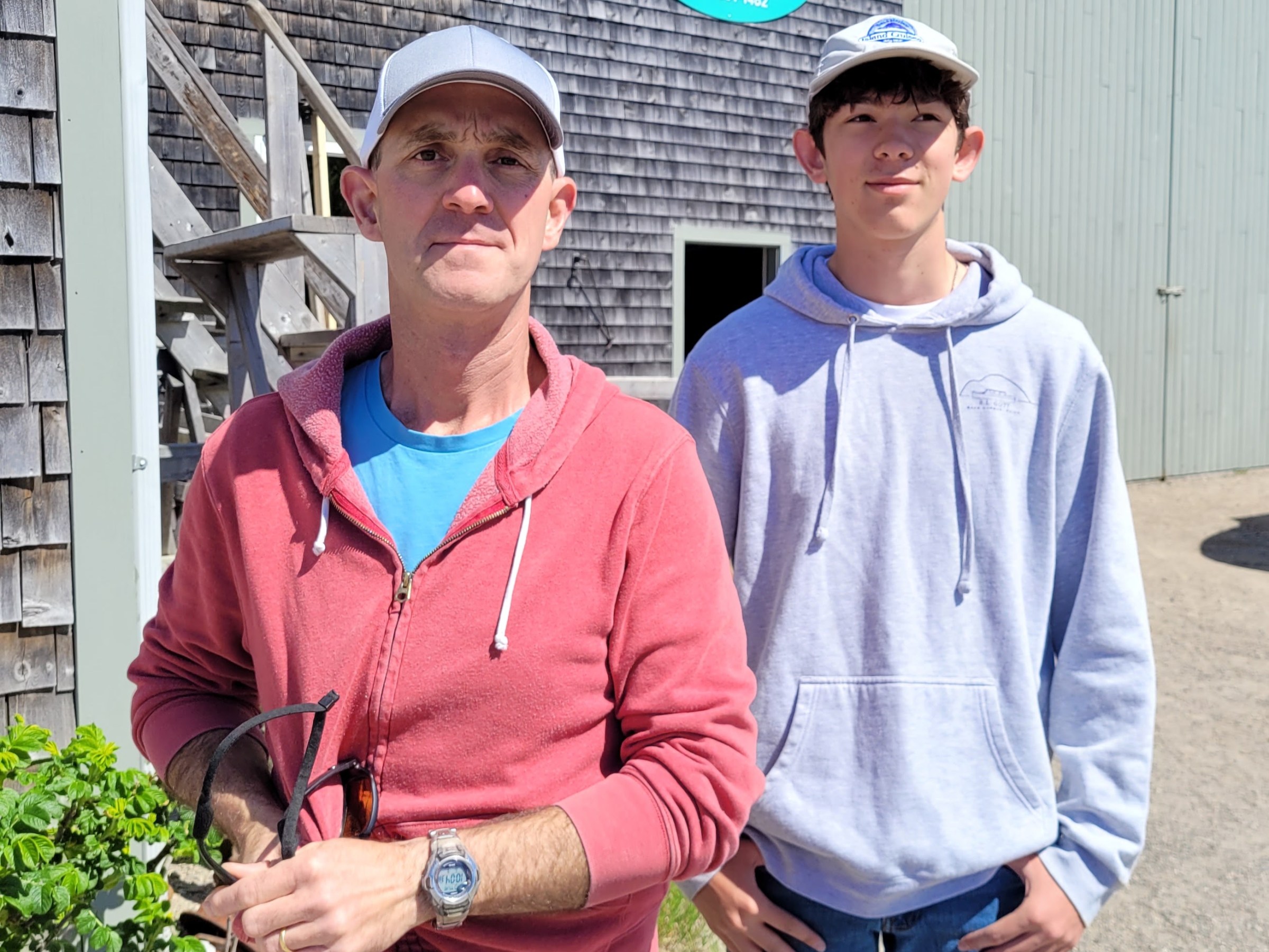 Two people standing outside a building with a marine sign, wearing casual clothes and caps.
