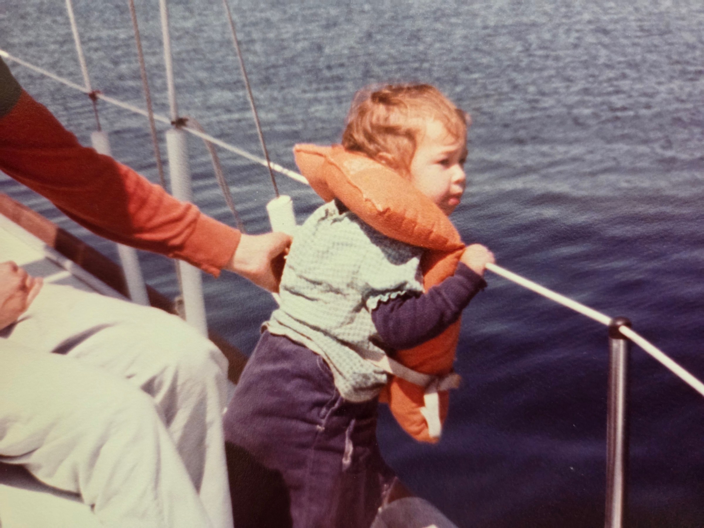 Toddler in life jacket on boat, held by adult near water.
