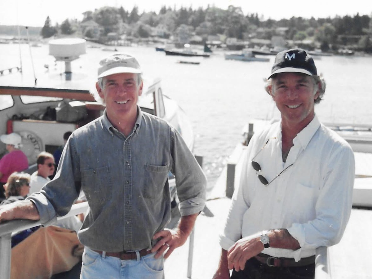 Two men in casual clothes and caps standing on a dock near boats.