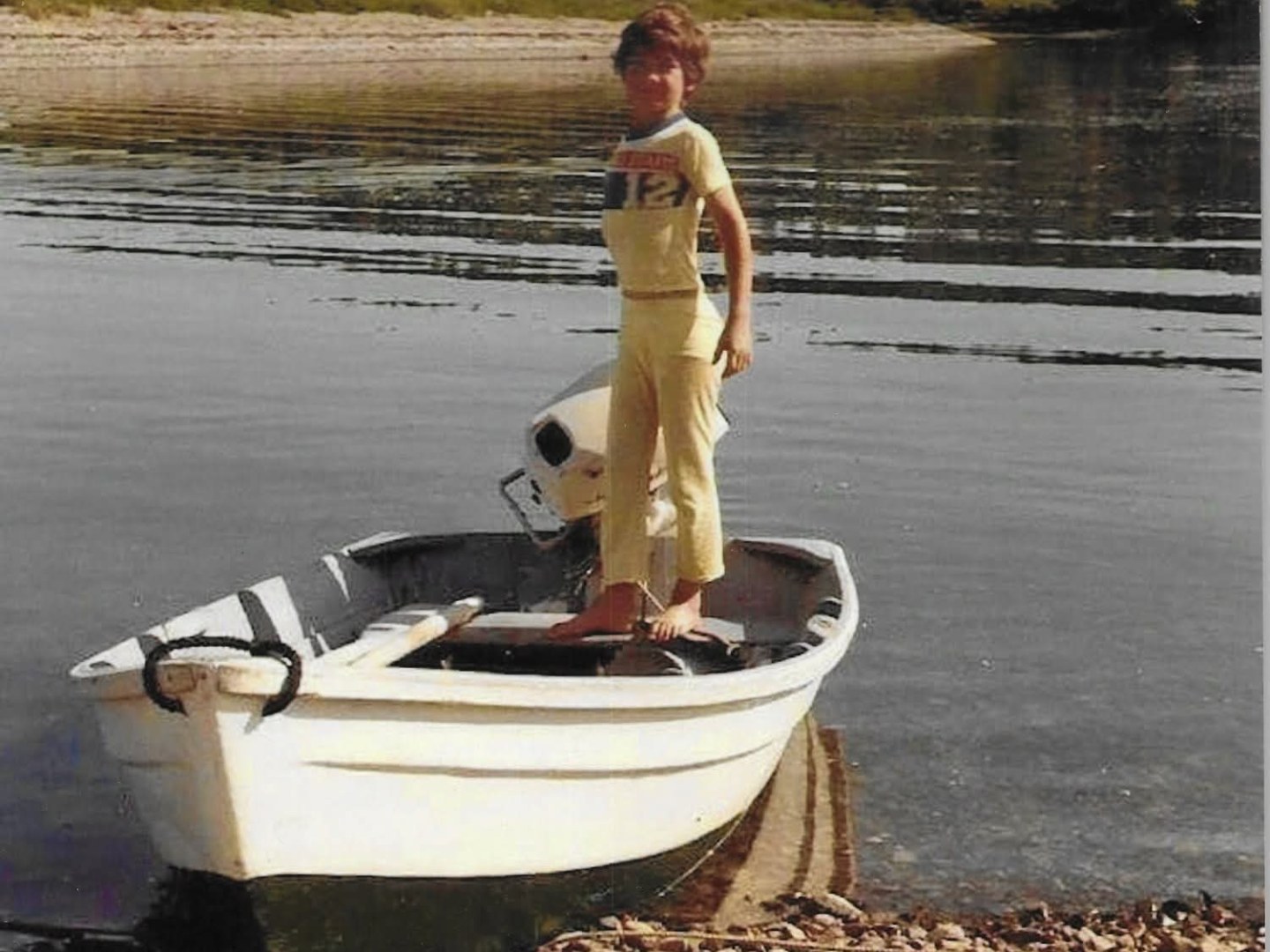 Person standing on a small boat by a rocky shore with forested background.