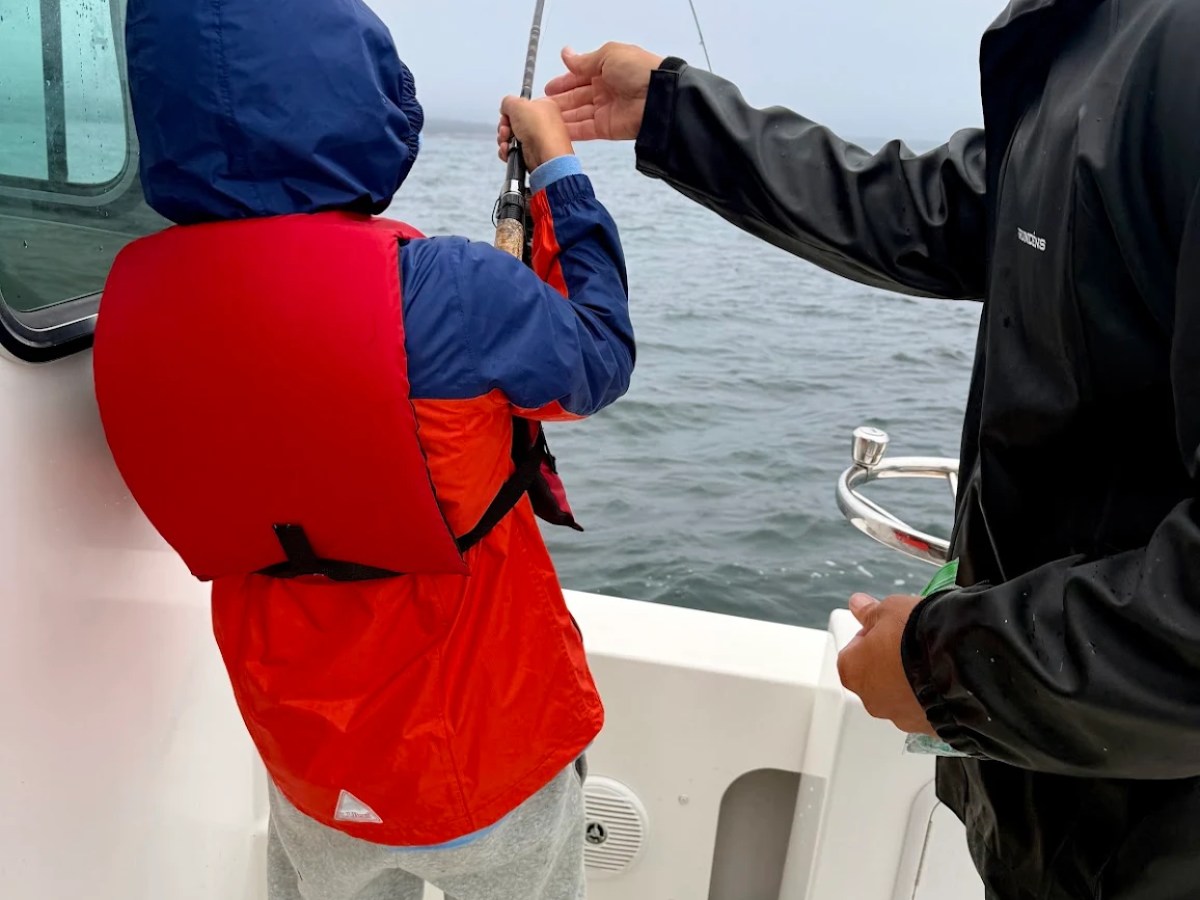 Two people fishing on a boat, one holding a fishing rod, both in rain jackets.