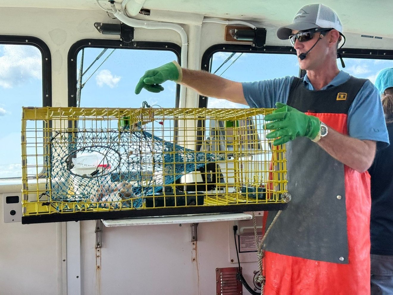 Person in gloves demonstrates a yellow lobster trap on a boat.