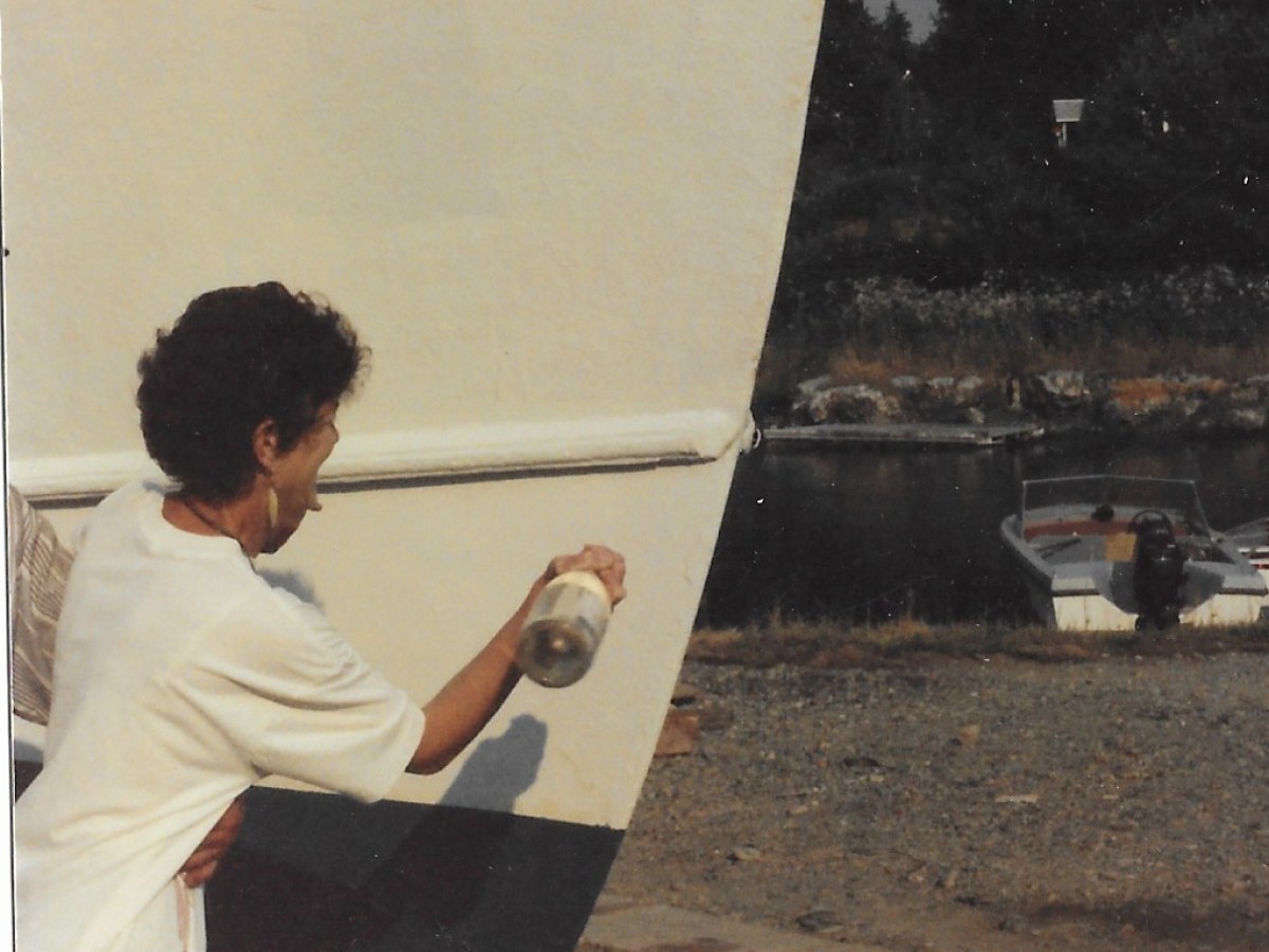 Person painting a boat hull on land near water and small boats.