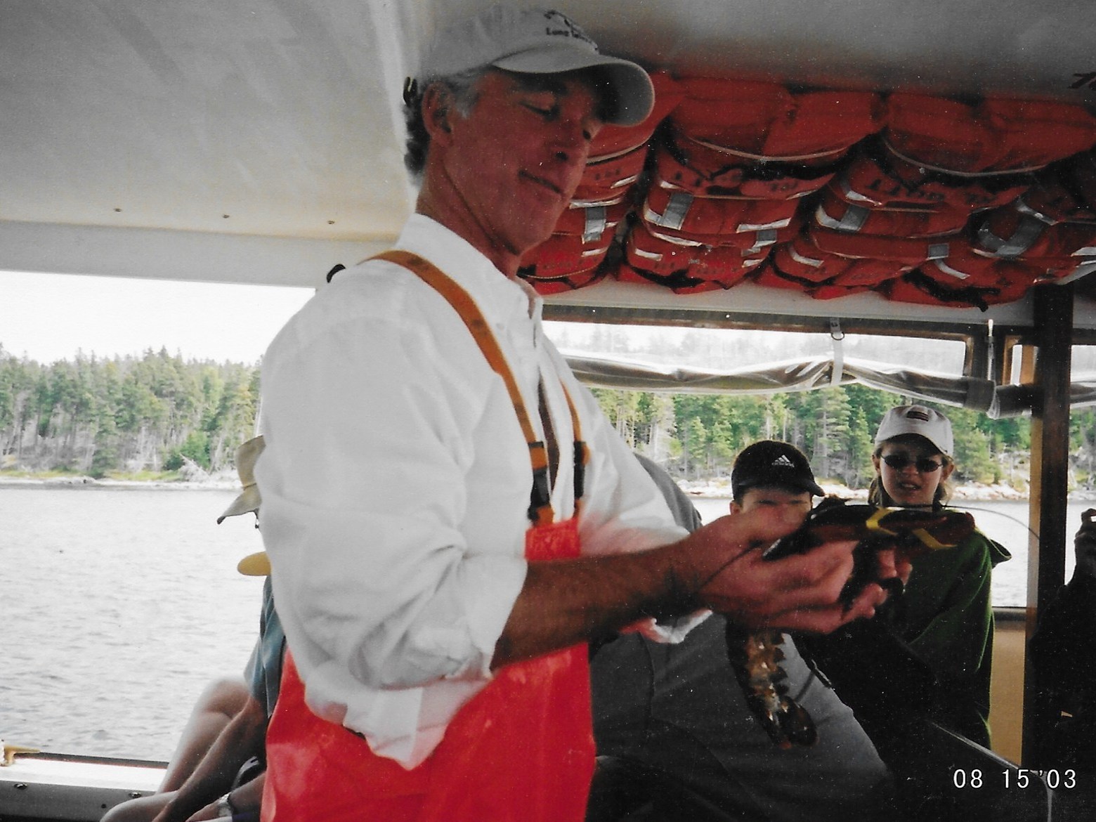 Man in red overalls holding lobster on boat with people and life jackets.