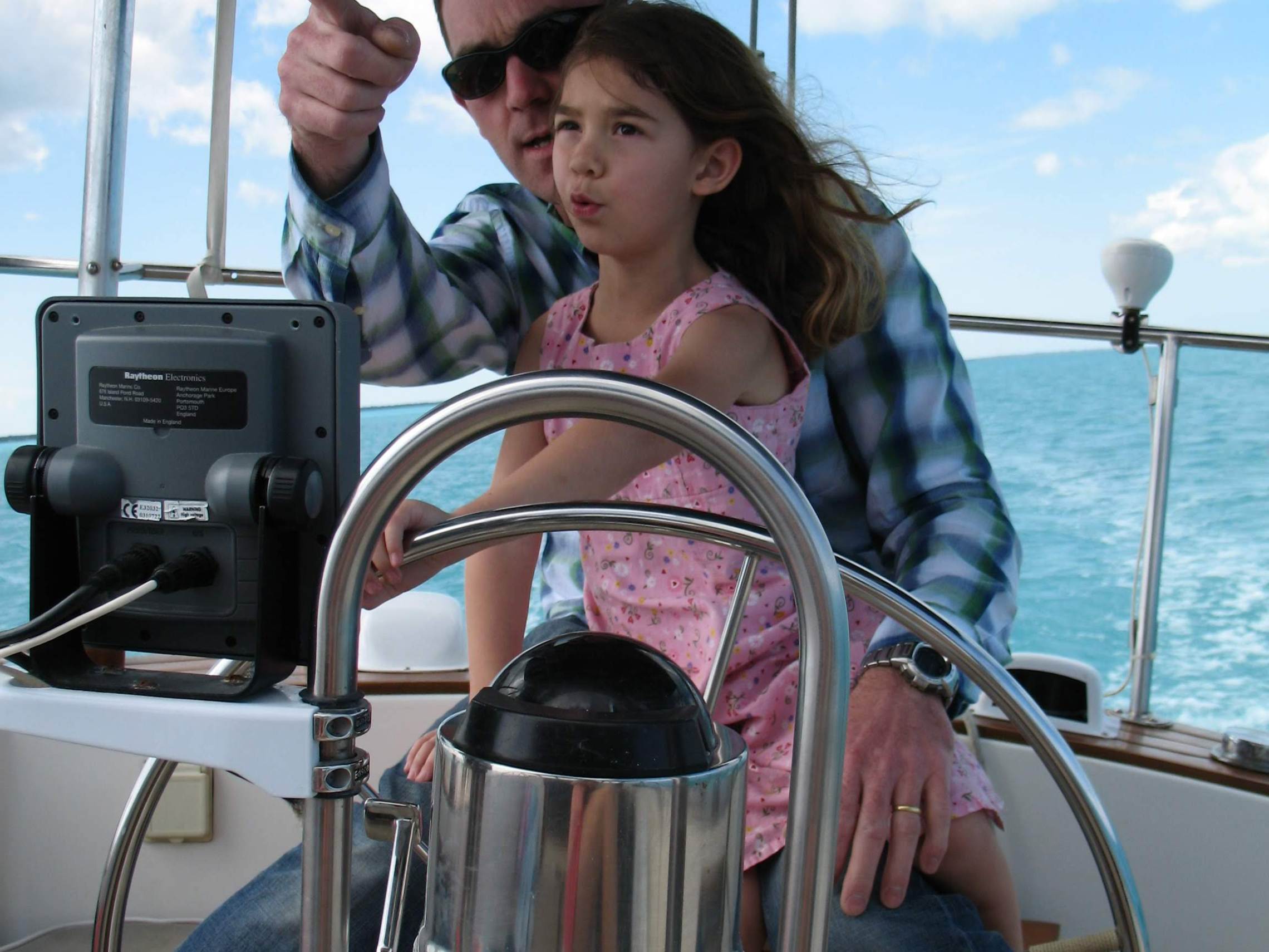 Man and girl steering a boat on a sunny day with ocean in background.